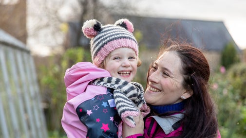 Young girl with stripey hat and scarf on, being held by a woman with long dark hair by a greenhouse.